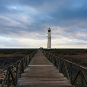 A picturesque wooden walkway leading to Faro de Morro Jable lighthouse under a cloudy sky in Spain.