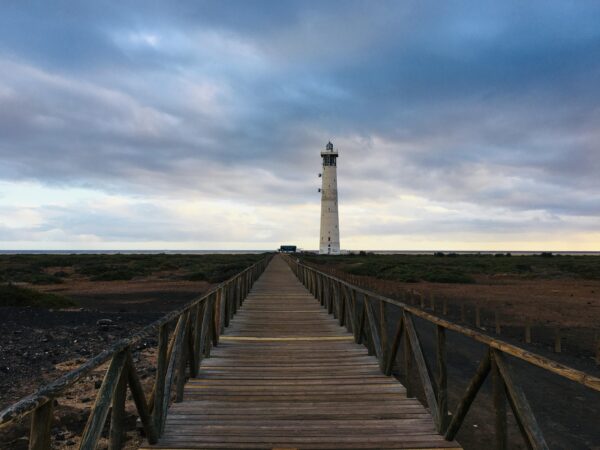 A picturesque wooden walkway leading to Faro de Morro Jable lighthouse under a cloudy sky in Spain.